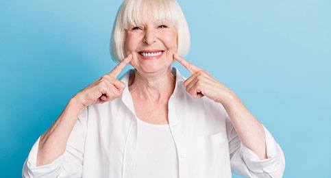 An older woman wearing white points to her healthy smile thanks to dental implants